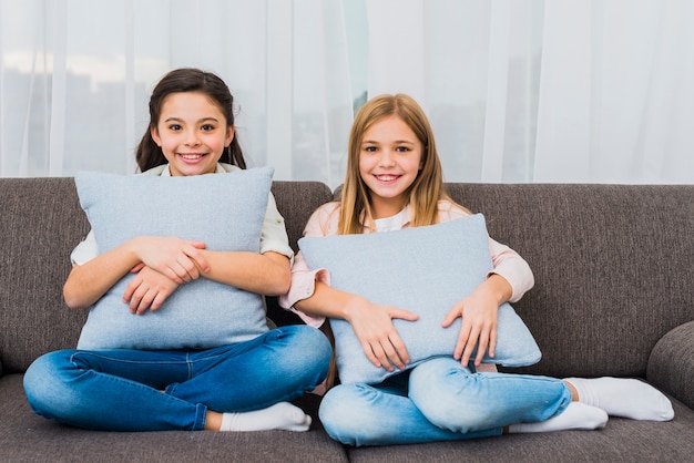 Retrato de dos muchachas sonrientes que se sientan en el sofá con los amortiguadores azules a disposición que miran a la cámara
