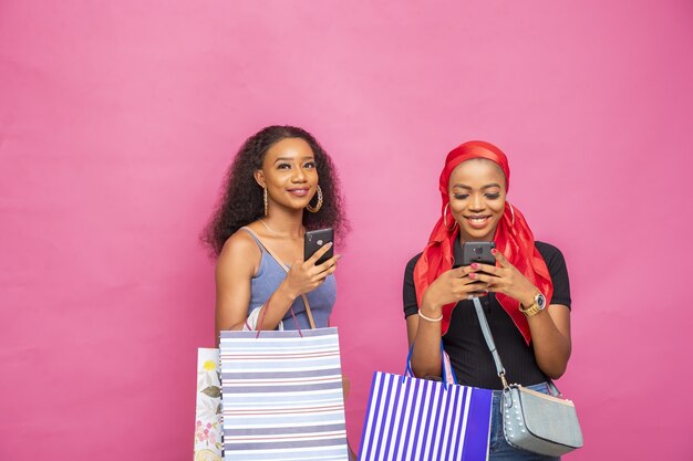 Retrato de dos mujeres africanas sosteniendo bolsas de la compra mientras usa sus teléfonos inteligentes