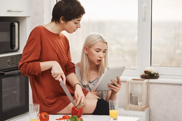 Retrato de dos mujeres atractivas que se sientan en cocina y que leen algo en tableta, expresando curiosidad e interés mientras preparan la ensalada. Las niñas pasan la prueba de lo bien que se conocen