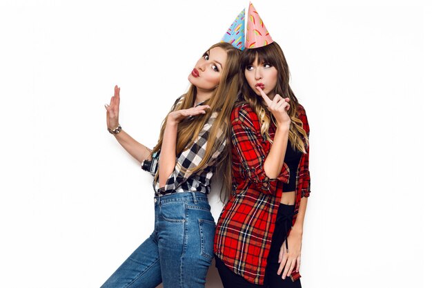 Retrato de dos mujeres bonitas posando en la pared blanca con sombreros de fiesta