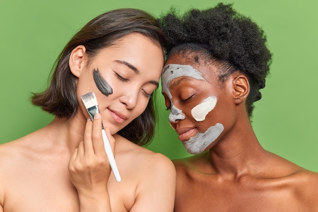 Retrato de dos mujeres diversas de pie sin camisa una al lado de la otra aplicar mascarilla peladora de belleza de arcilla con cepillo cosmético se ocupa de la pose de la piel contra la pared verde del estudio