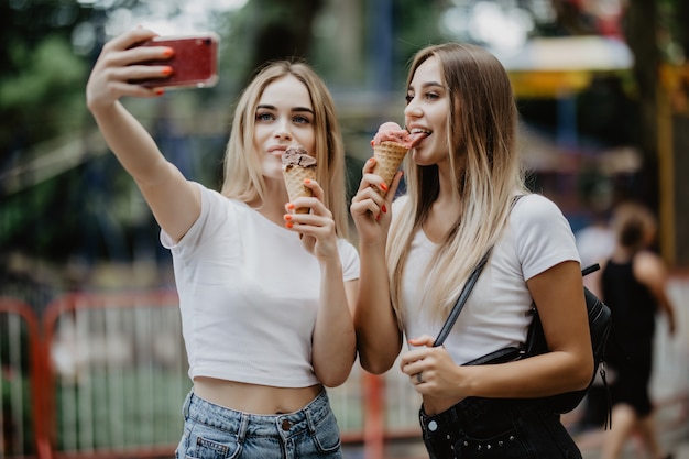 Retrato de dos mujeres jóvenes de pie juntos comiendo helado y tomando selfie en la calle del verano.