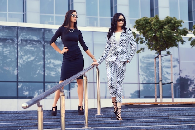 Retrato de dos mujeres de negocios vestidas con ropa formal elegante, de pie en los escalones en un centro posando contra el fondo de un rascacielos.