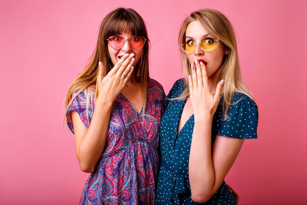 retrato de dos mujeres positivas mejores amigas divirtiéndose en la pared rosa, vistiendo vestidos de verano vintage estampados brillantes y gafas de sol, chismeando juntas, emociones emocionadas.