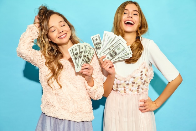 Retrato de dos mujeres rubias encantadas felices vistiendo ropa de verano regocijándose ganar y sosteniendo dinero en efectivo aislado sobre la pared azul