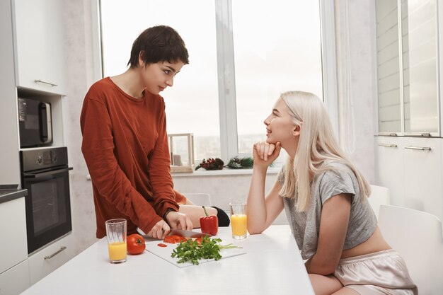 Retrato de dos mujeres sentadas en la cocina, beber jugo y hacer ensalada mientras habla y hace bromas por la mañana. Chica rubia coquetea con su novia mientras prepara el desayuno