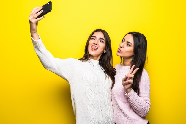 Retrato de dos niñas alegres vestidas con suéteres de pie y tomando un selfie aislado sobre pared amarilla