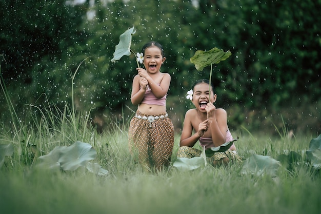 Retrato de dos niñas encantadoras en la tradición tailandesa se visten y se ponen una flor hermosa en la oreja, se quitan la hoja de loto Protegen las gotas de lluvia con un espacio de copia alegre