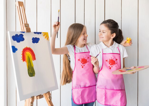 Retrato de dos niñas sonrientes en un delantal rosa burlándose mientras pinta en el lienzo