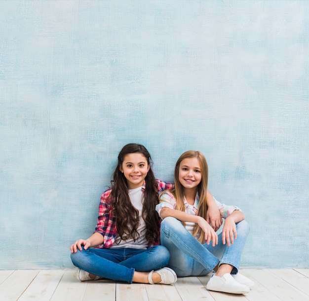 Retrato de dos niñas sonrientes sentados juntos contra la pared azul pintada