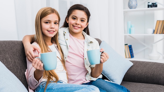 Retrato de dos niños femeninos sentados juntos en el sofá sosteniendo tazas de café en la mano