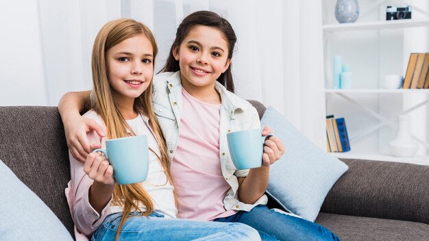 Retrato de dos niños femeninos sentados juntos en el sofá sosteniendo tazas de café en la mano