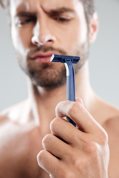 Retrato de un dudoso hombre barbudo concentrado mirando maquinilla de afeitar