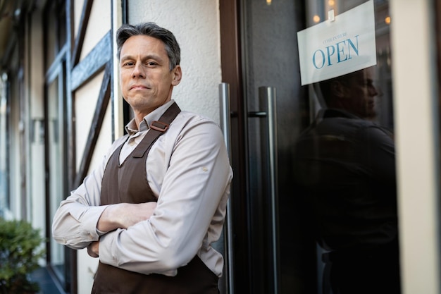Retrato del dueño del bar con los brazos cruzados parados en la puerta de entrada y mirando la cámara.