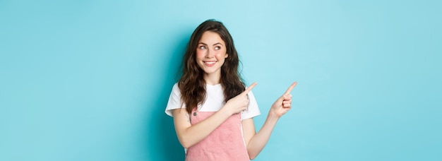 Foto gratuita retrato de una dulce y alegre niña con peto rosa y camiseta blanca mirando el producto señalando con el dedo