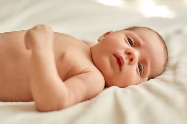 Retrato de dulce bebé recién nacido acostado en la cama sobre una manta blanca, estudiando cosas externas, encantador niño lindo, hermoso niño mirando a otro lado.