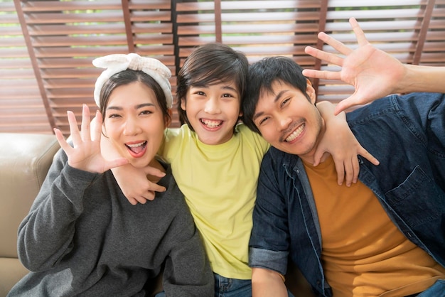 Retrato de una dulce familia asiática sentada y sonriendo en la sala de estar, el hijo abraza a su padre con la mano amorosa, toma el brazo del padre y la madre por detrás y mira la cámara con felicidad y alegría en casa