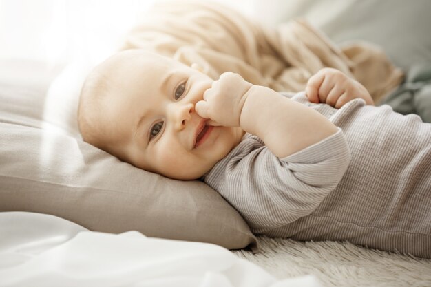 Retrato de dulce hija recién nacida sonriente acostado en la cama acogedora. El niño mira la cámara y toca la cara con sus pequeñas manos. Momentos de la infancia.
