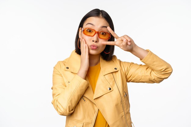 Retrato de una elegante chica asiática moderna con gafas de sol y una chaqueta amarilla que muestra un gesto de paz frente a un rostro sonriente feliz de fondo blanco