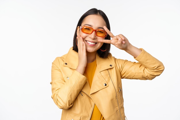 Retrato de una elegante chica asiática moderna con gafas de sol y una chaqueta amarilla que muestra un gesto de paz frente a un rostro sonriente feliz de fondo blanco