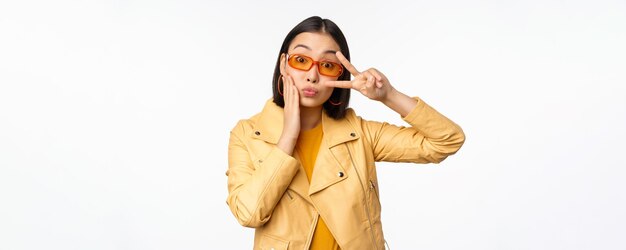 Retrato de una elegante chica asiática moderna con gafas de sol y una chaqueta amarilla que muestra un gesto de paz frente a un rostro sonriente feliz de fondo blanco