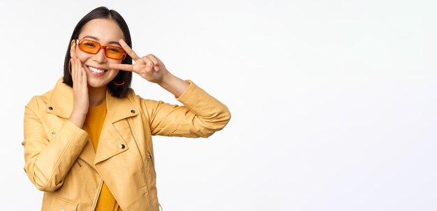 Retrato de una elegante chica asiática moderna con gafas de sol y una chaqueta amarilla que muestra un gesto de paz frente a un rostro sonriente feliz de fondo blanco