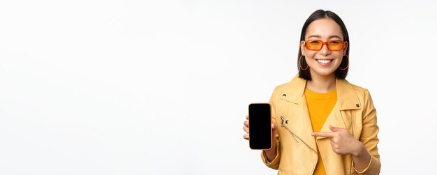Retrato de una elegante chica coreana con gafas de sol sonriendo señalando con el dedo a la pantalla del teléfono inteligente que muestra una aplicación de teléfono móvil sobre fondo blanco