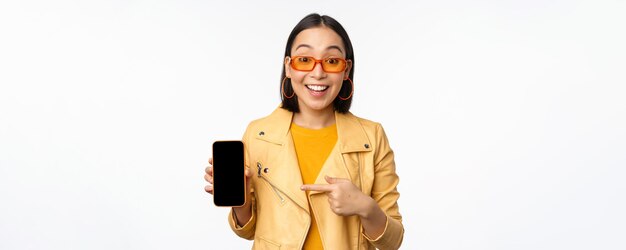 Retrato de una elegante chica coreana con gafas de sol sonriendo señalando con el dedo a la pantalla del teléfono inteligente que muestra m