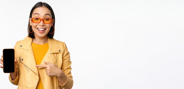 Retrato de una elegante chica coreana con gafas de sol sonriendo señalando con el dedo a la pantalla del teléfono inteligente que muestra m