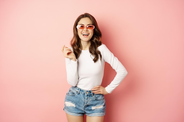 Retrato de una elegante chica glam con gafas de sol riendo y sonriendo de pie sobre un fondo rosa de vacaciones