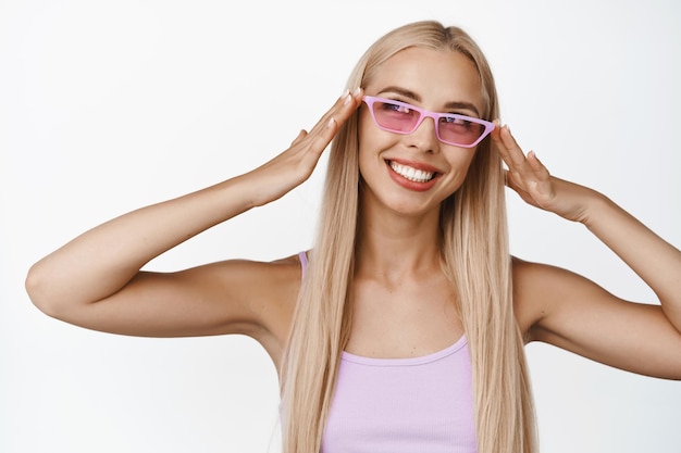 Retrato de una elegante chica rubia con gafas de sol rosas sonriendo feliz y mirando a un lado con una expresión facial despreocupada sobre fondo blanco