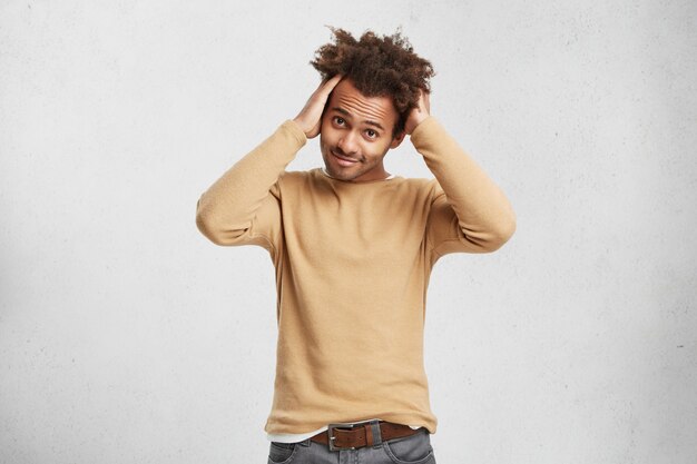 Retrato de elegante hombre de piel oscura con pelo rizado, viste jeans y suéter, mantiene las manos en la cabeza