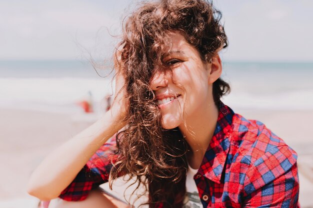 Retrato de elegante jovencita con cabello oscuro ondulado y hermosos ojos sentada junto al mar a la luz del sol. Verano, recreación, vacaciones, emociones felices.