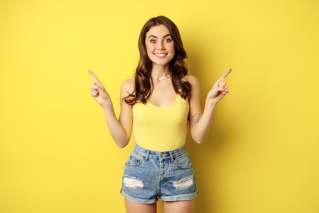 Retrato de una elegante modelo femenina joven, mostrando publicidad, apuntando al logo o pancarta, demostrando dos formas, opciones en la tienda, de pie sobre fondo amarillo.