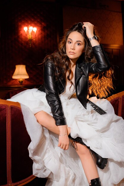 Retrato de una elegante modelo femenina con el pelo largo y rizado vestido con un vestido de novia blanco, una chaqueta de cuero negro y botas sentadas en un sofá en una habitación oscura posando seriamente y mirando a la cámara