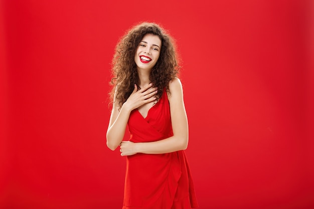 Retrato de elegante mujer caucásica encantadora complacida y agradecida con peinado rizado sosteniendo la palma en el pecho sonriendo y riendo divertido posando en elegante vestido de noche sobre fondo rojo.
