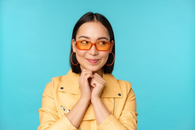 Retrato de una elegante mujer coreana con gafas de sol sonriendo y mirando a un lado el logo coqueto de pie sobre fondo azul Copiar espacio