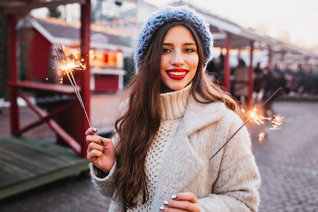 Retrato de elegante mujer de pelo castaño con elegante bata blanca con luces de Bengala. Foto al aire libre de la romántica chica europea en boina azul posando con luces de bengala en la ciudad de desenfoque