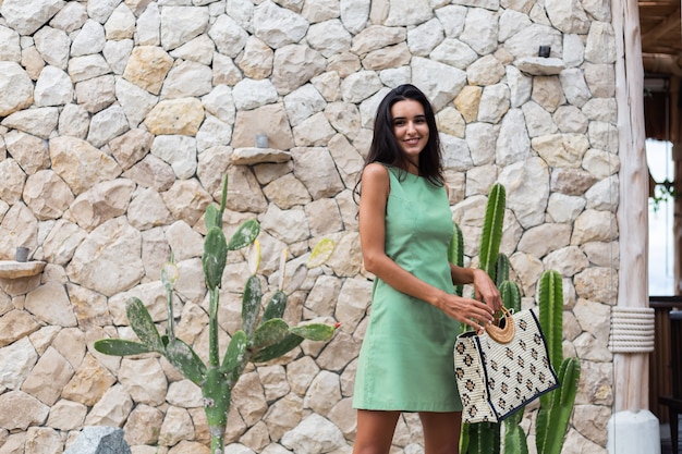 Retrato de elegante mujer sonriente linda feliz en elegante vestido verde de verano con bolsa con sombrero de paja sobre fondo de pared de piedra blanca y cactus