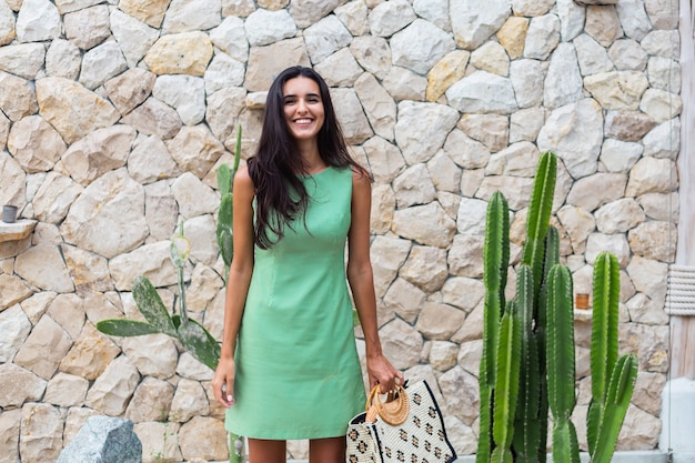 Retrato de elegante mujer sonriente linda feliz en elegante vestido verde de verano con bolsa con sombrero de paja sobre fondo de pared de piedra blanca y cactus