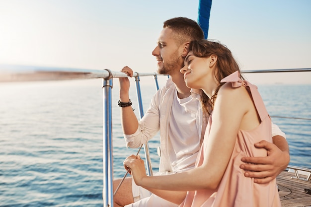 Retrato de la elegante pareja de enamorados, abrazándose mientras está sentado en la proa del yate privado y disfrutando de la vista del mar. El esposo llevó a su esposa a un hermoso país cálido, celebrando la luna de miel