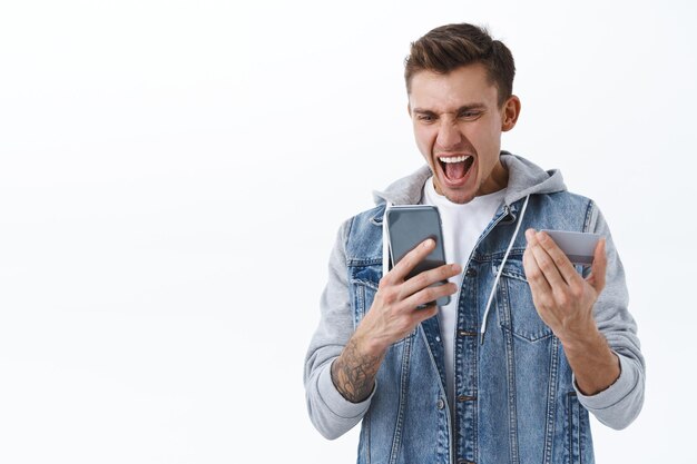 Retrato de emotivo chico caucásico rubio gritando en el teléfono móvil como comprobar su cuenta bancaria vacía