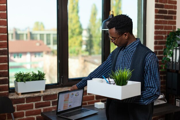 Retrato de un empleado afroamericano que fue despedido cerrando una computadora portátil con datos comerciales mientras sostenía una bandeja con pertenencias personales antes de irse. Despedido del escritorio de limpieza del trabajador de inicio antes de irse a casa.