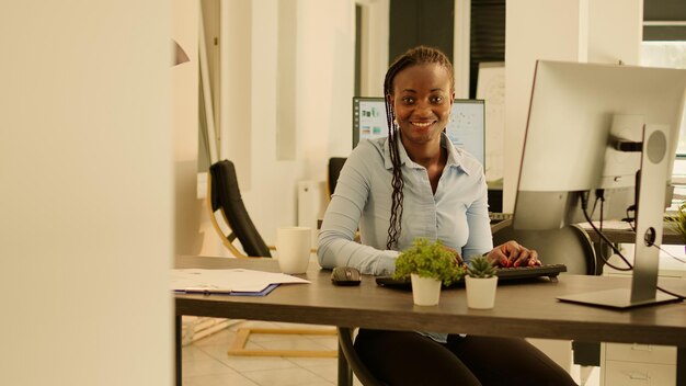 Retrato de un empleado afroamericano que trabaja en un informe comercial, usando una computadora en el escritorio de la oficina. Tomar notas y enviar correos electrónicos durante la puesta del sol, generar ideas en el trabajo de inicio.