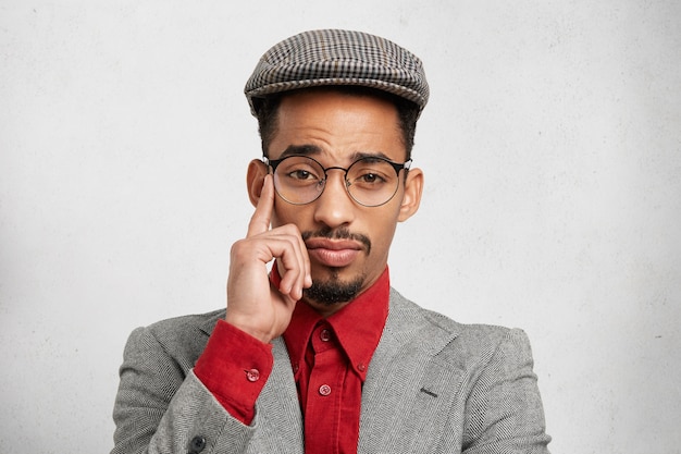 Retrato de empleado masculino de raza mixta pensativo pensativo viste gorra de moda, camisa roja y chaqueta, tiene expresión sabia