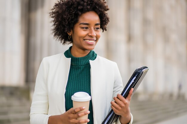 Retrato de empresaria afro sosteniendo una taza de café y un portapapeles mientras camina al aire libre en la calle. Concepto urbano y empresarial.