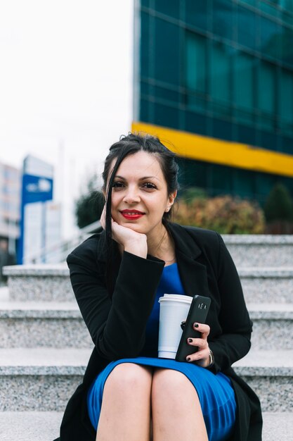 Retrato de una empresaria feliz con la taza de la disposición y el teléfono móvil que se sientan en escalera