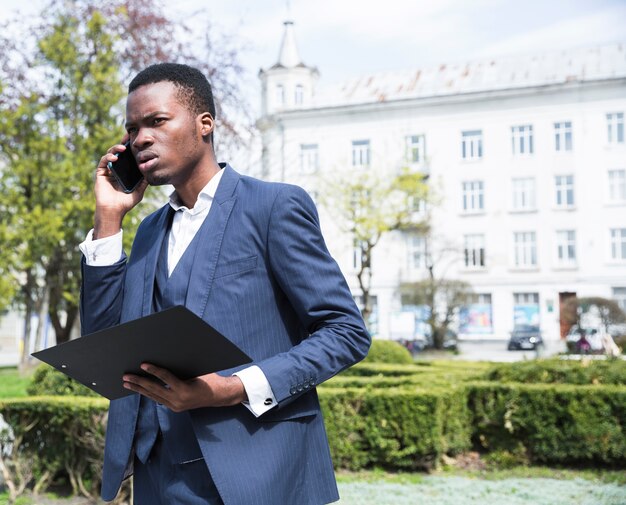 Retrato de una empresaria joven africana sosteniendo portapapeles hablando por teléfono móvil