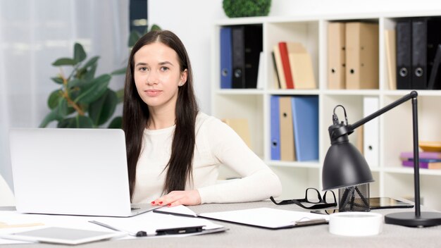 Retrato de una empresaria joven con la computadora portátil en el escritorio que mira a la cámara