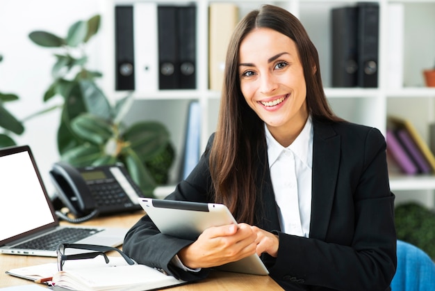 Retrato de una empresaria joven que se sienta en el escritorio que sostiene la tableta digital en su mano